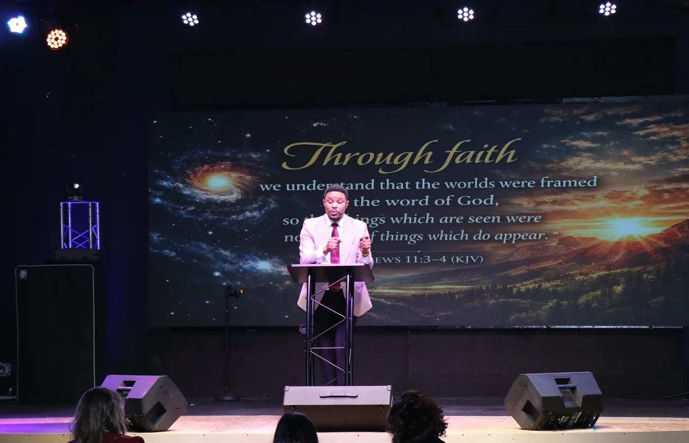 A large church auditorium with stage lighting and a wall-sized LED screen showing Hebrews 11 verses 3 to 4, with the speaker at a podium in front of it.