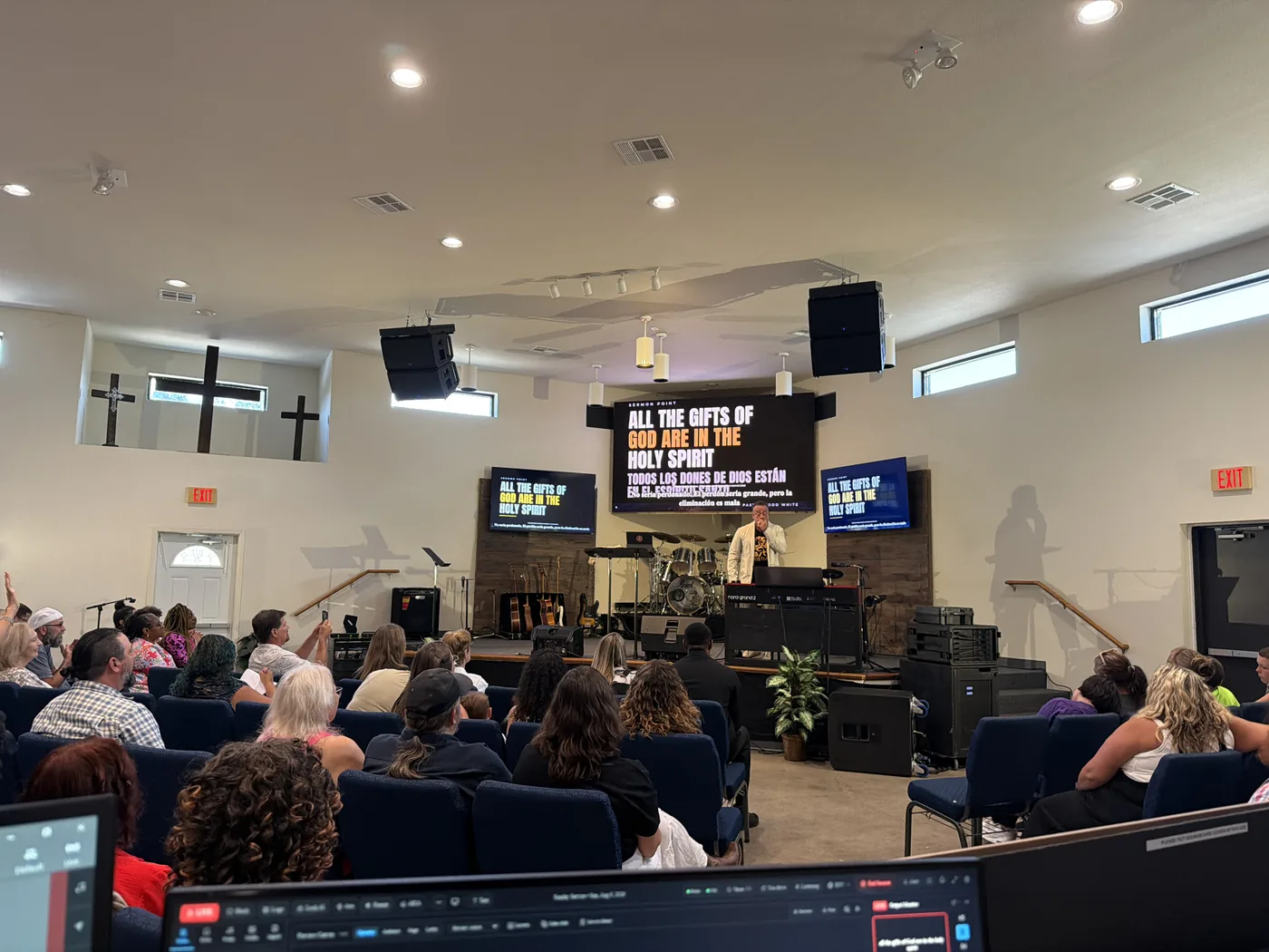 A church auditorium photographed from the back row. The congregation faces three screens carrying a sermon point in English and Spanish, and in the foreground sits the laptop running Aria, showing the service timeline.