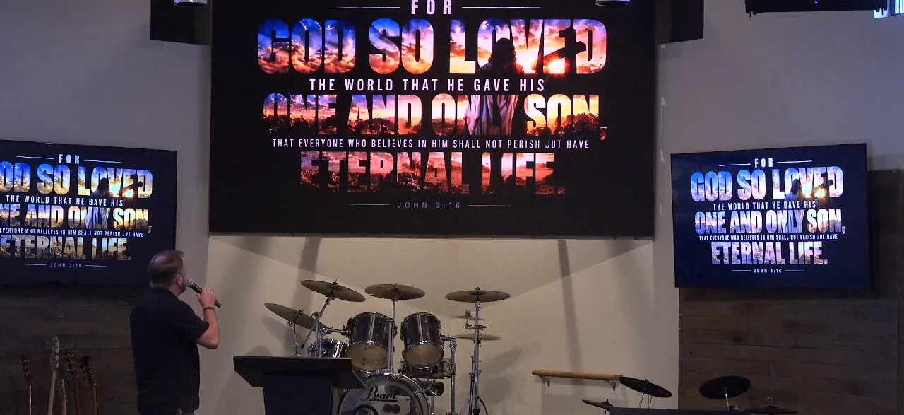 John 3:16 artwork filling the main screen and both side screens of a church auditorium, while a man stands at the podium speaking into a microphone.