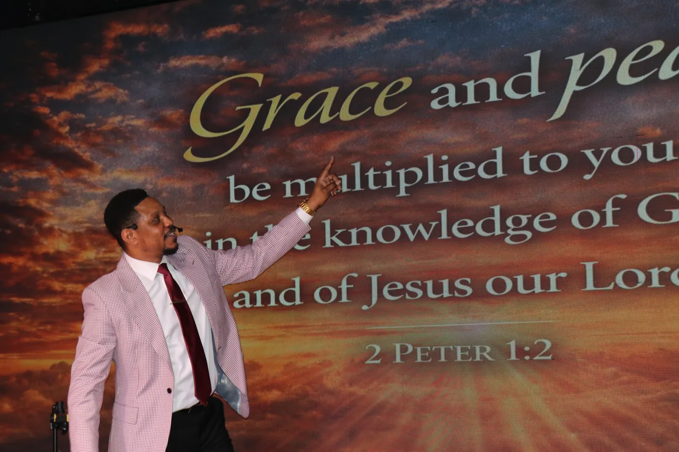 A speaker on stage turned towards a wall-sized screen, pointing up at 2 Peter 1 verse 2 — 'Grace and peace be multiplied to you in the knowledge of God and of Jesus our Lord' — set in large type over a sunset sky.