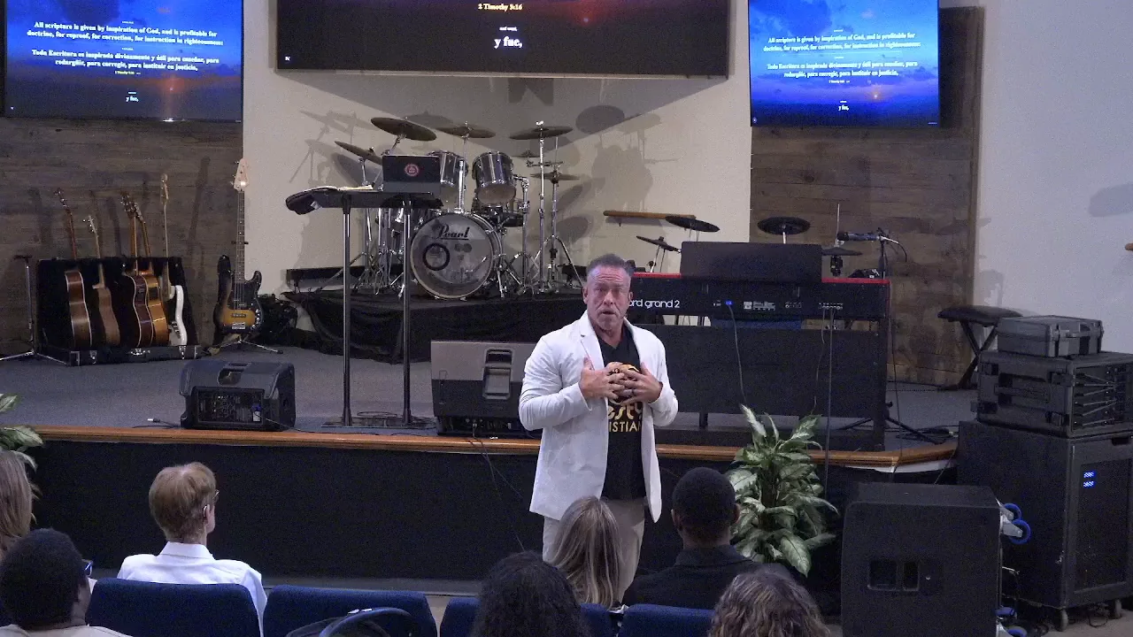 The same service from the room's own camera: the speaker mid-sentence, with the screens behind and beside him showing 2 Timothy 3 verse 16 in English and Spanish.